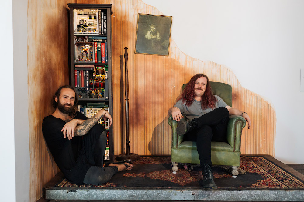 Jackson Harvey and Alex Towler sit in the corner of a room, Harvey on the floor, which is covered in a Persian rug, and Towler in a vintage lime green leather armchair. There is a narrow bookshelf in one corner, a lampstand with no lamp and a black and white portrait hanging on the wall.