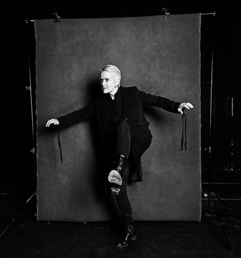 Sally Richardson stands in front of a photographer's backdrop. She has one leg raised and bent, as though she is about to take a step over an obstacle, and her arms are stretched out for balance. The image is black and white.