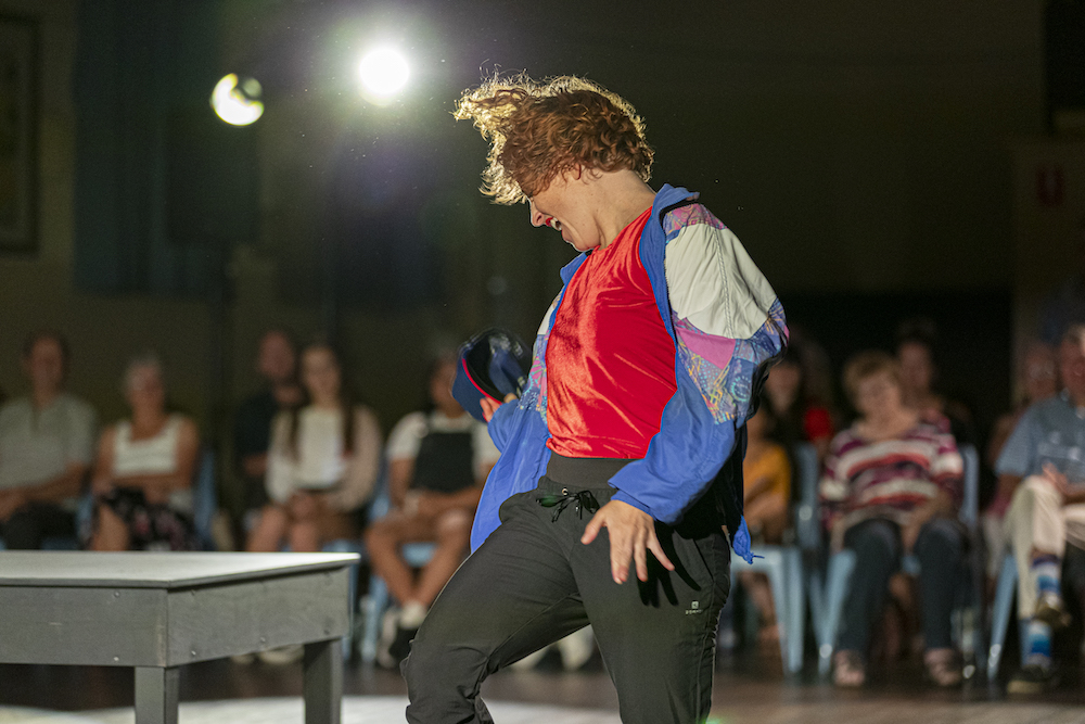 Natalie Allen is captured mid-action - she's twisting slightly to the left of the frame and her hair flairs slightly with the movement. Although her arms are close to her sides, her fingers are extended and she looks like she might be calling something out. She wears a gaudy parasilk trackie top over a red shirt and black pants.