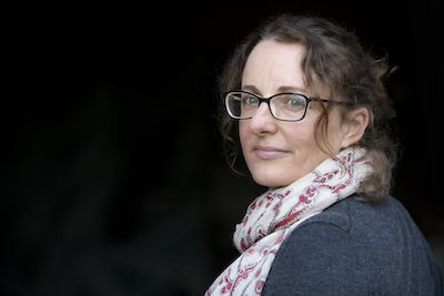 A headshot of Zoe Deleuil. She is positioned at right angles to the camera and turns her head to the lens. She has wavy hair tied back, with some strands left out to frame her face. She wears glasses, a white scarf with a red pattern and a grey top.